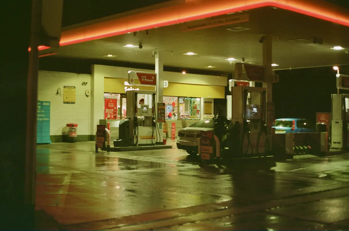 A nighttime photo of a wet Shell gas station with red neon lighting, showing fuel pumps, a convenience store, and cars at…