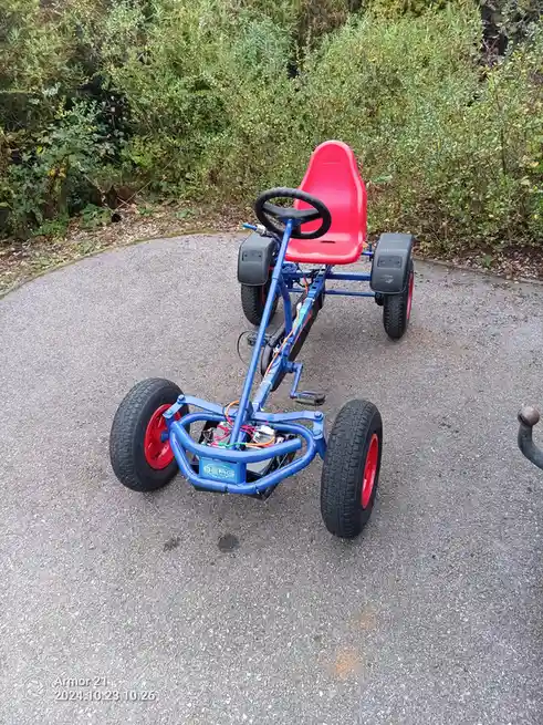 A blue and red go-kart with exposed wiring and a battery pack sits on a paved path, surrounded by green bushes. The overlaid…