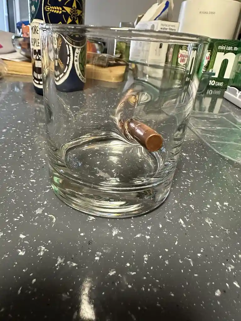 A clear, empty rocks glass sits on a speckled countertop with a single copper-colored bullet resting inside it. The…