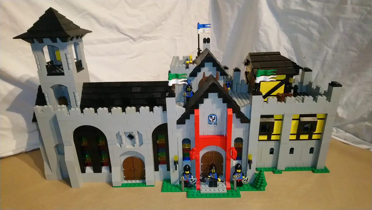 A detailed Lego castle model with grey stone walls, black roofs, and multiple towers, featuring small Lego knights in blue…