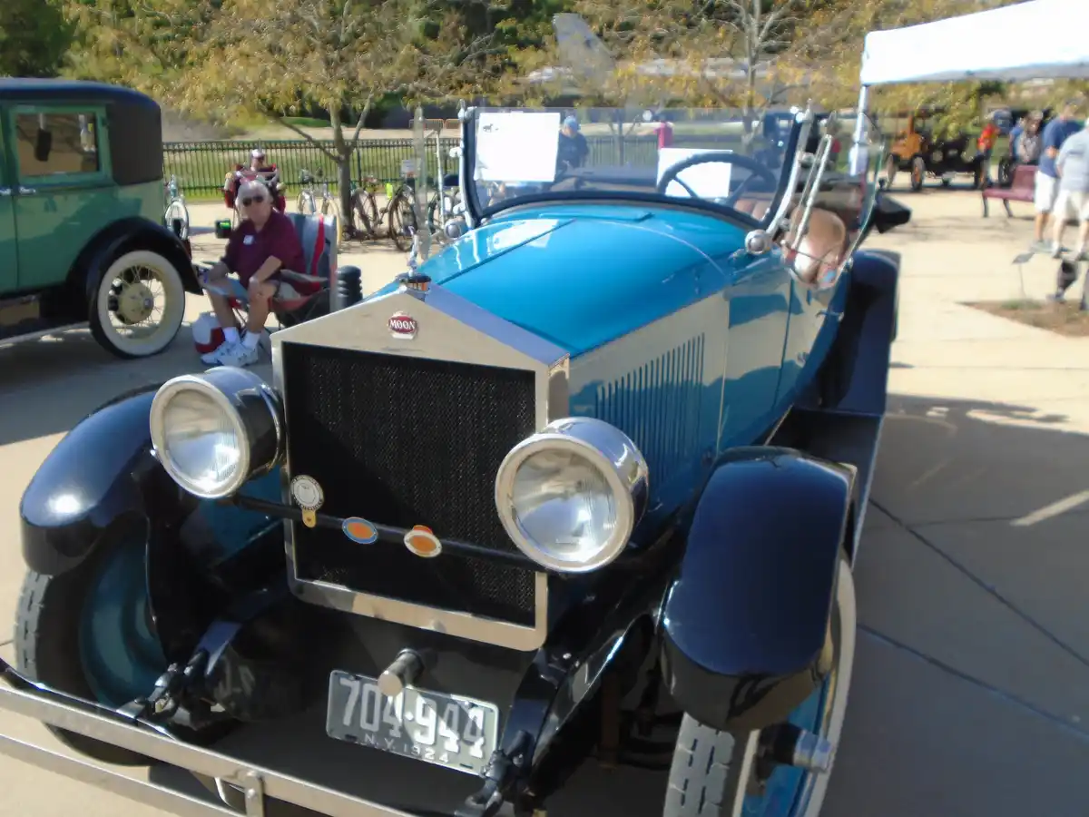 A vintage blue and black Moon automobile from 1924 is parked at an outdoor car show, with a man in a maroon shirt and…