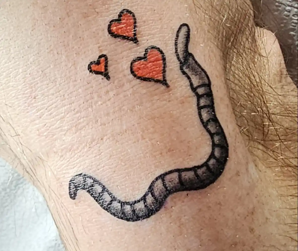 A close-up photo shows a tattoo on human skin depicting a segmented worm with three small red hearts floating above it. The…