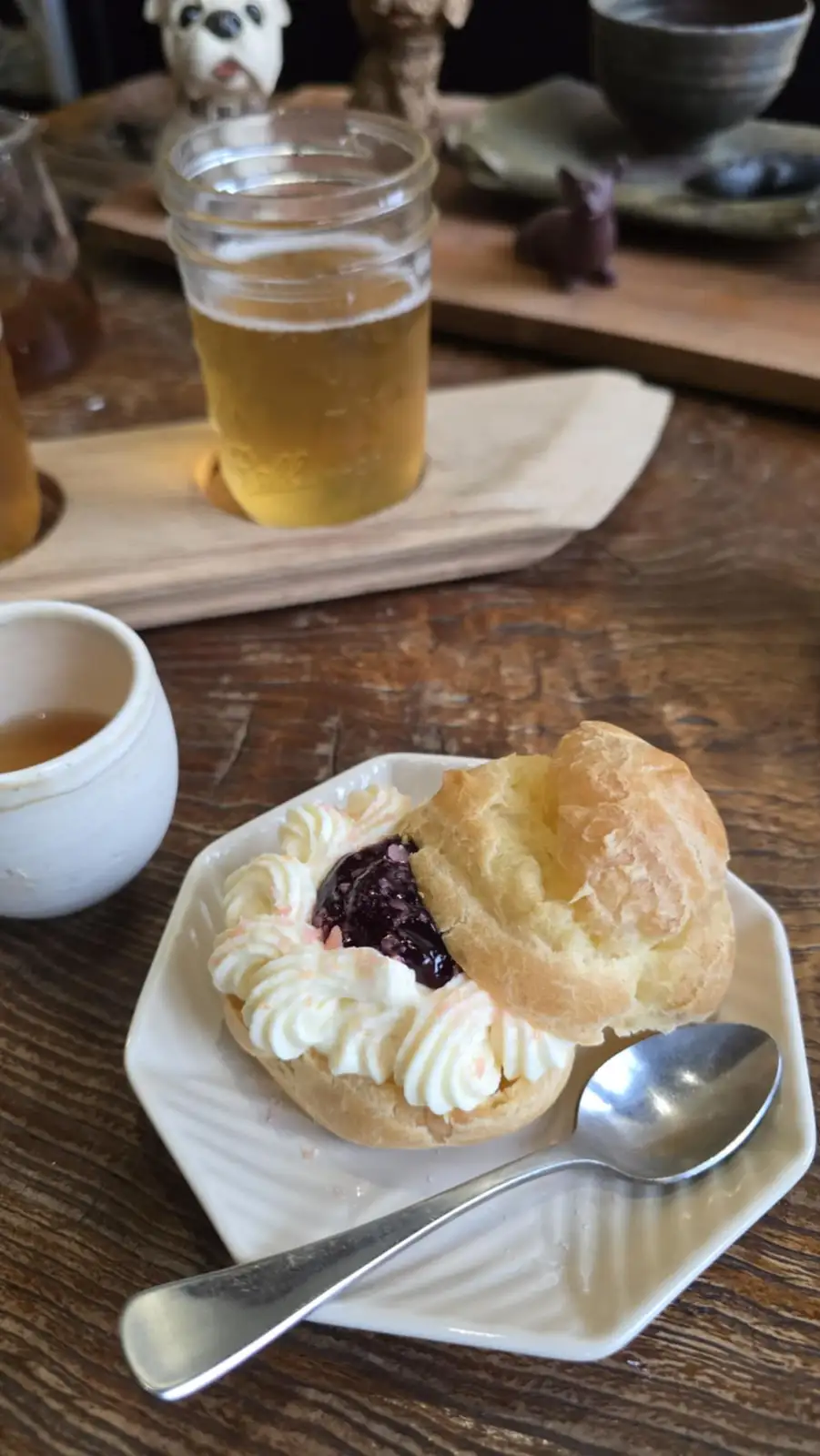 a close-up photo shows a cream puff split open on a white plate, filled with dark jam and piped whipped cream, with a metal…