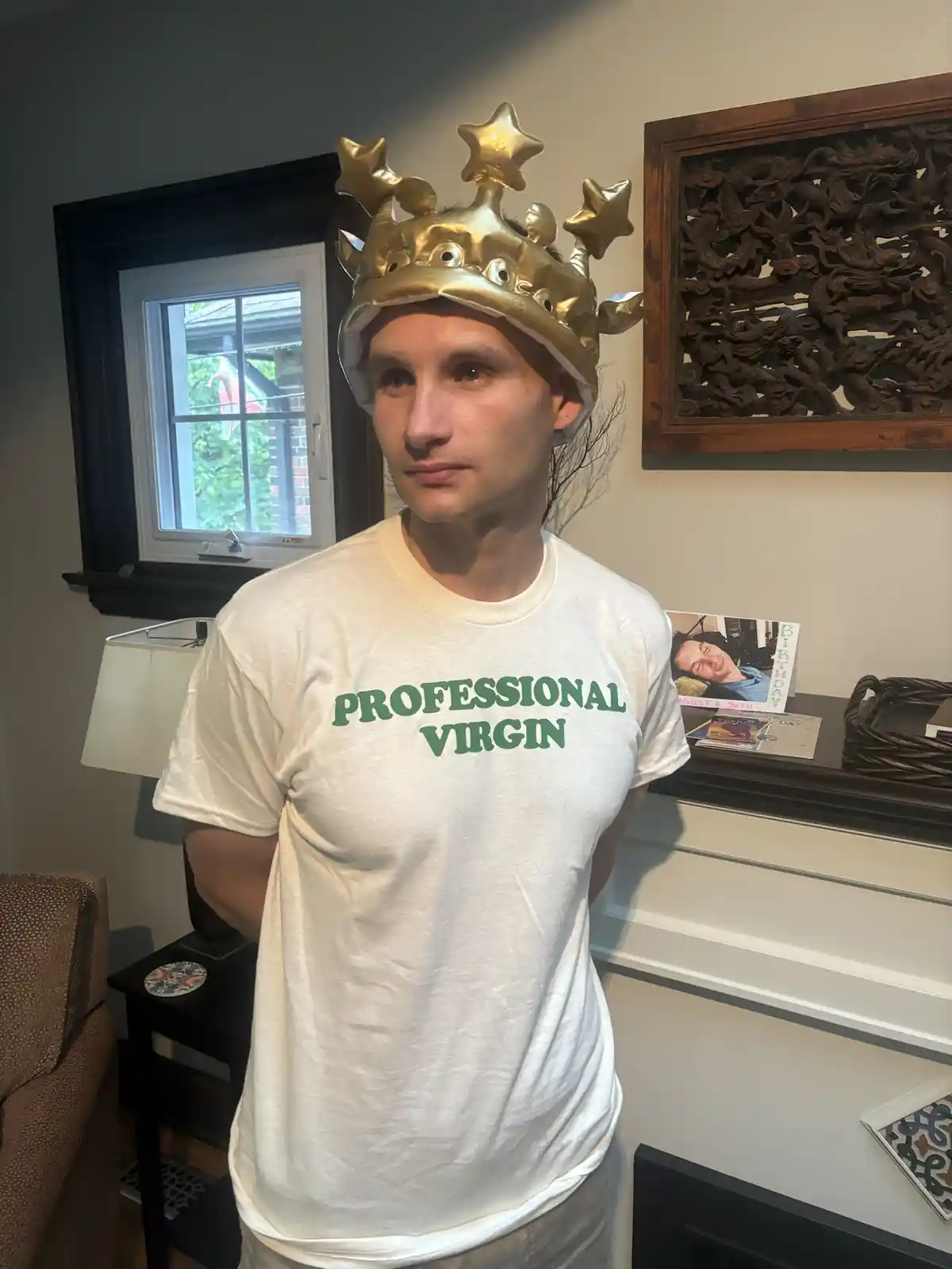 A person stands indoors wearing a shiny gold crown with star-shaped points and a white t-shirt that reads 'PROFESSIONAL…