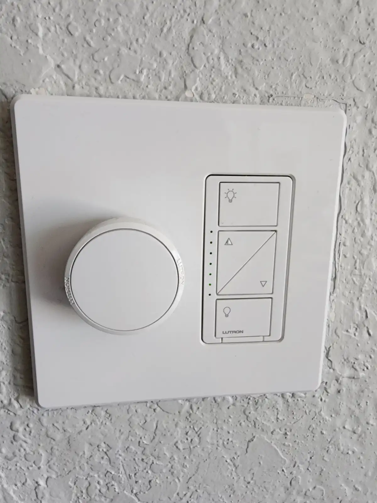 A white Lutron smart light switch is mounted on a textured wall, featuring a large circular dial on the left and a vertical…