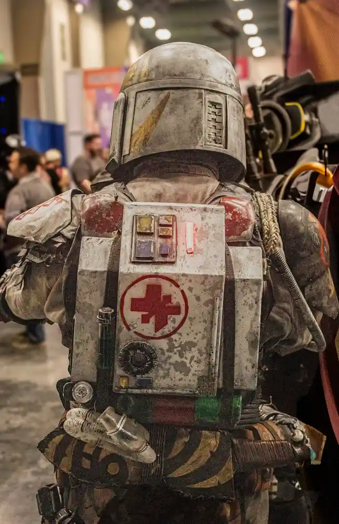 A cosplayer in a heavily weathered Mandalorian armor costume, viewed from behind, wears a large, worn medical pack with a…
