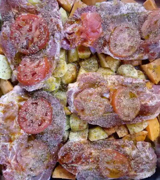 A close-up photo shows raw meat pieces arranged over chopped vegetables, including sweet potatoes and onions, all seasoned…