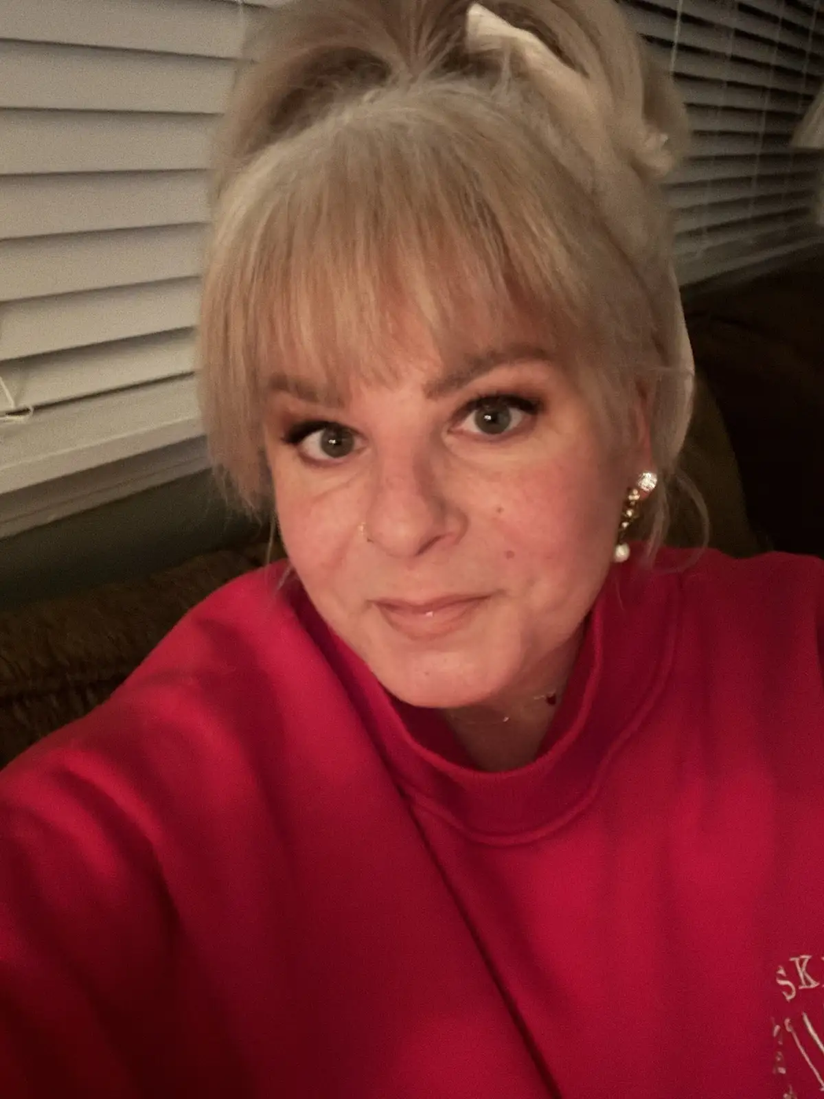 A selfie of a woman with blonde hair styled in a messy bun, wearing a red turtleneck sweater and pearl earrings, looking…