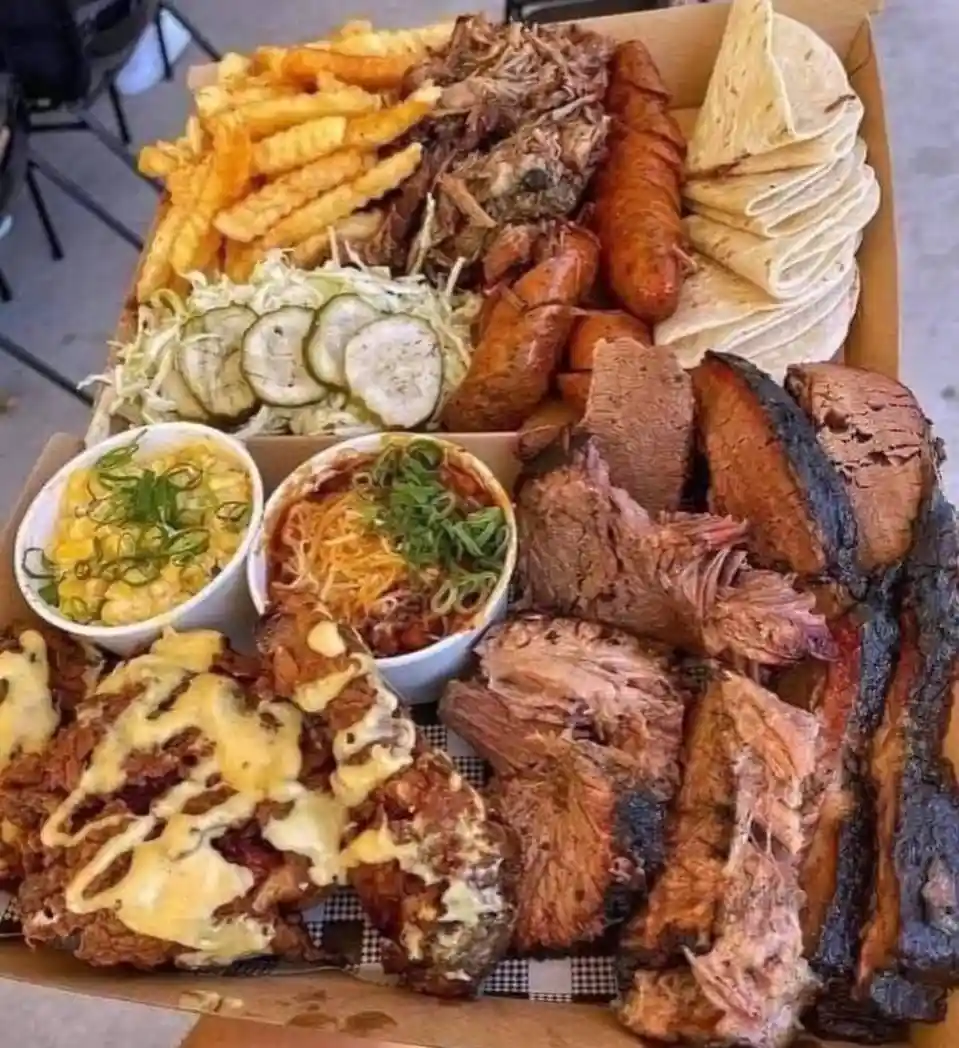 A large, overflowing cardboard box filled with an assortment of barbecue foods including pulled pork, smoked brisket, ribs…