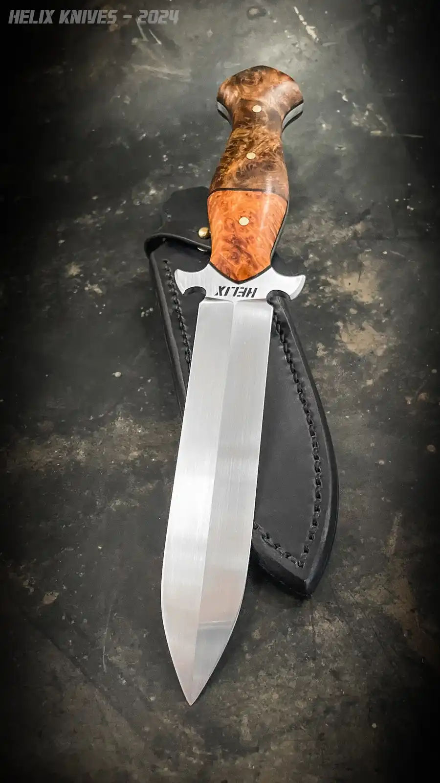 A close-up photo of a Helix brand knife with a wooden handle and silver blade, resting in a black leather sheath on a dark…