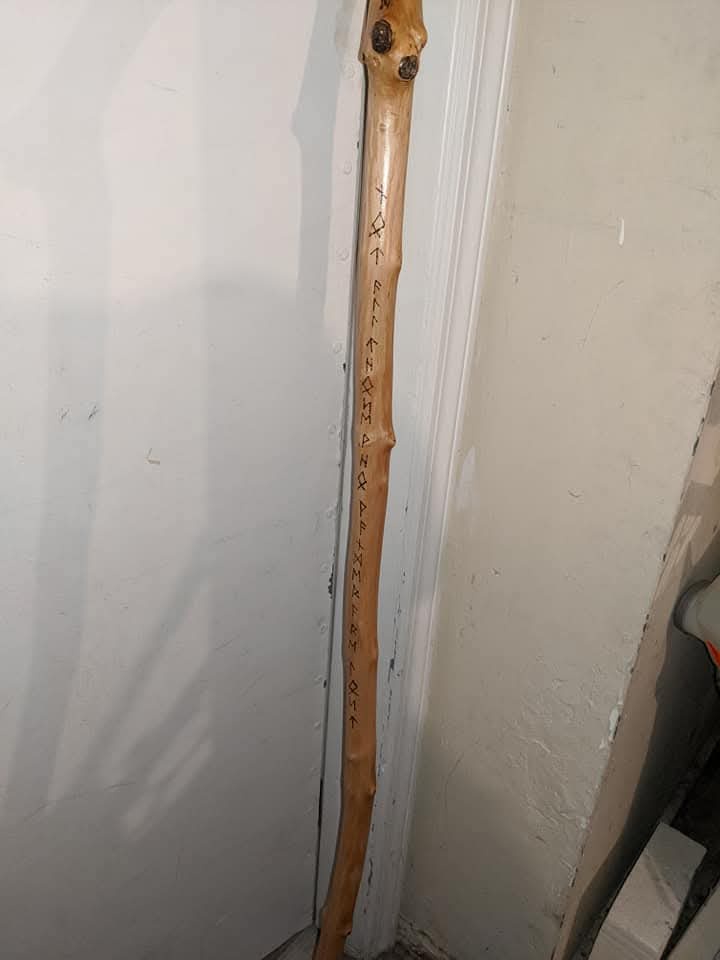A wooden walking stick leaning against a white wall has text carved vertically along its length, reading 'i'm not a wizard…