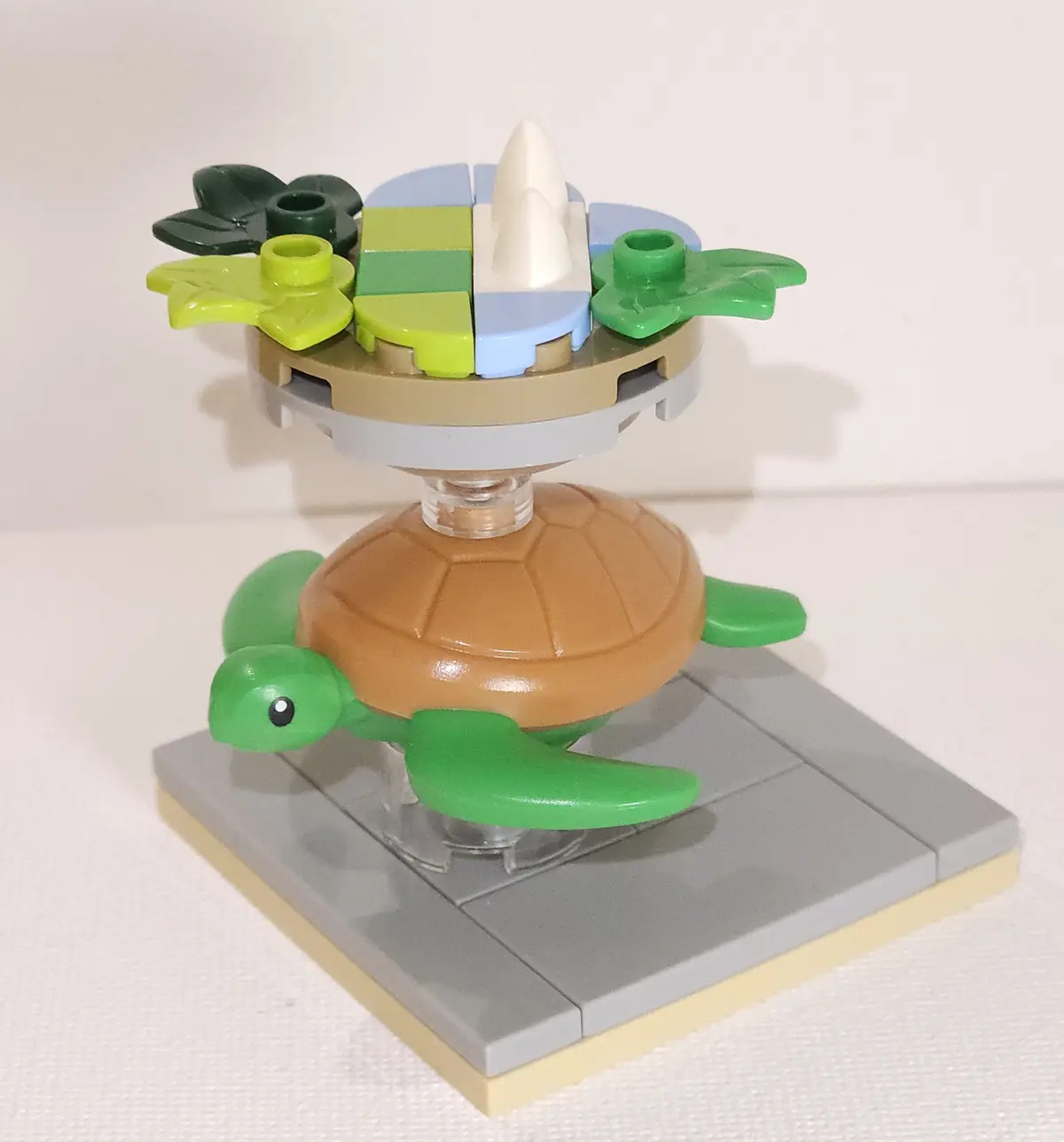 A LEGO model of a green turtle with a brown shell stands on a gray baseplate. On its back is a circular platform holding a…
