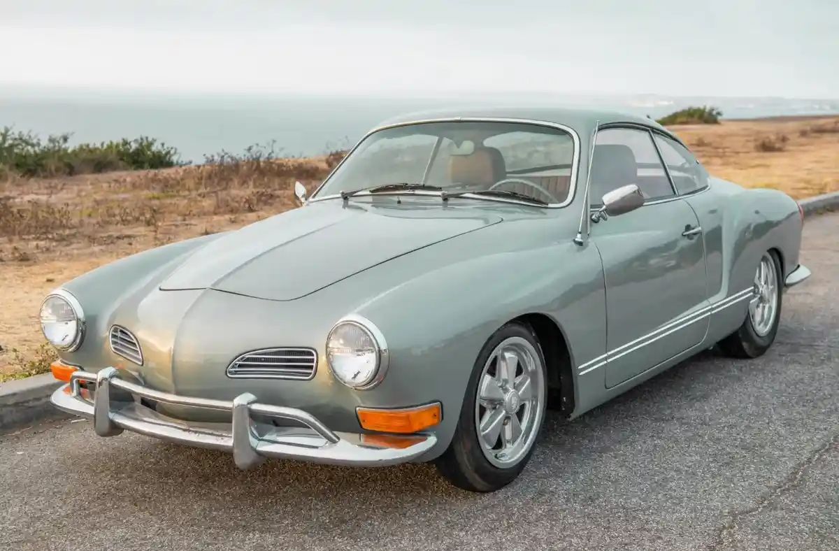 A light green vintage Volkswagen Karmann Ghia coupe is parked on an asphalt road overlooking a hazy ocean. The car has…