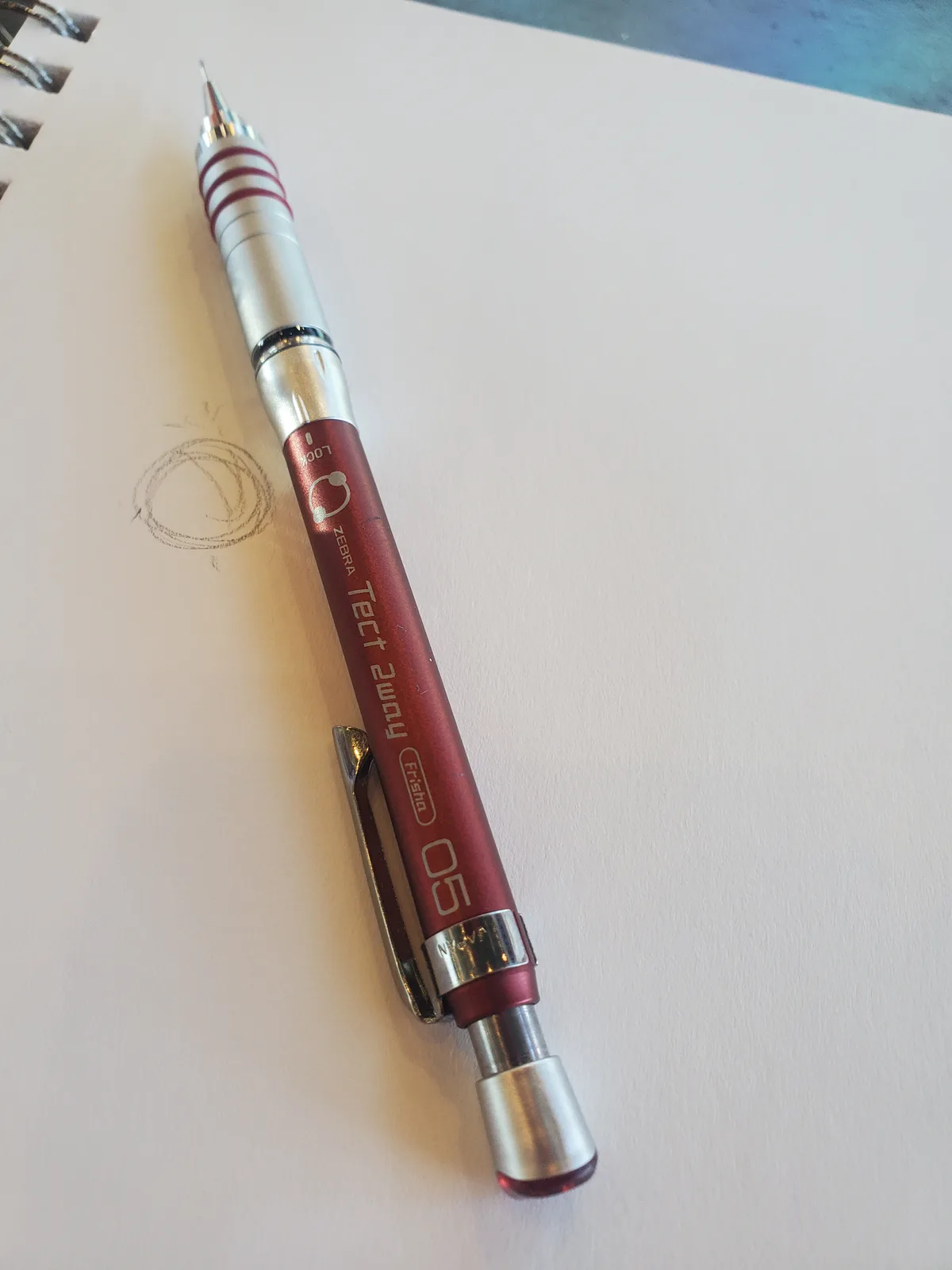 A red and silver Zebra Tect 2way mechanical pencil lies diagonally on a white spiral-bound notebook page. Beside the pencil…