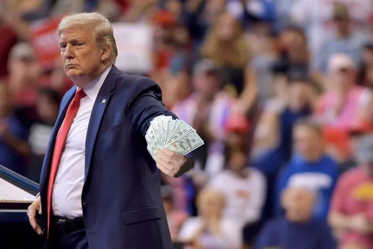 a man in a dark suit and red tie stands at a podium, holding a fan of US dollar bills in his right hand while looking away…