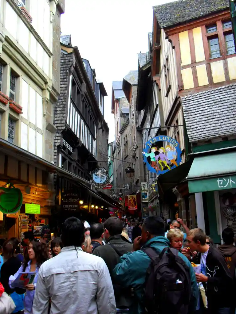 A crowded, narrow European street lined with half-timbered buildings and shop signs, including one reading 'LES LUTINS' with…