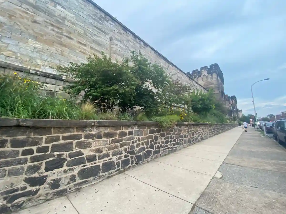 A photo shows a long, high stone wall with crenellations resembling a medieval castle, running alongside a modern sidewalk…