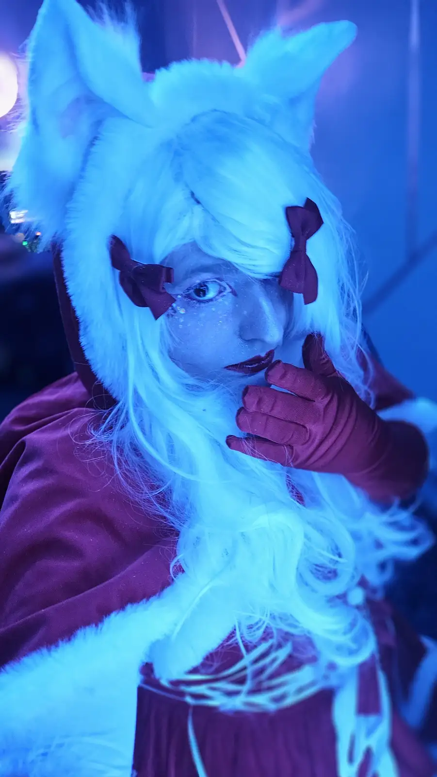A person in a blue-lit environment wears a white furry fox-eared hood, a white wig with dark red bows, and a dark red outfit…