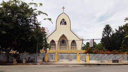 A beige church with a pointed steeple and cross sits behind a white fence, surrounded by trees and a paved road. The…
