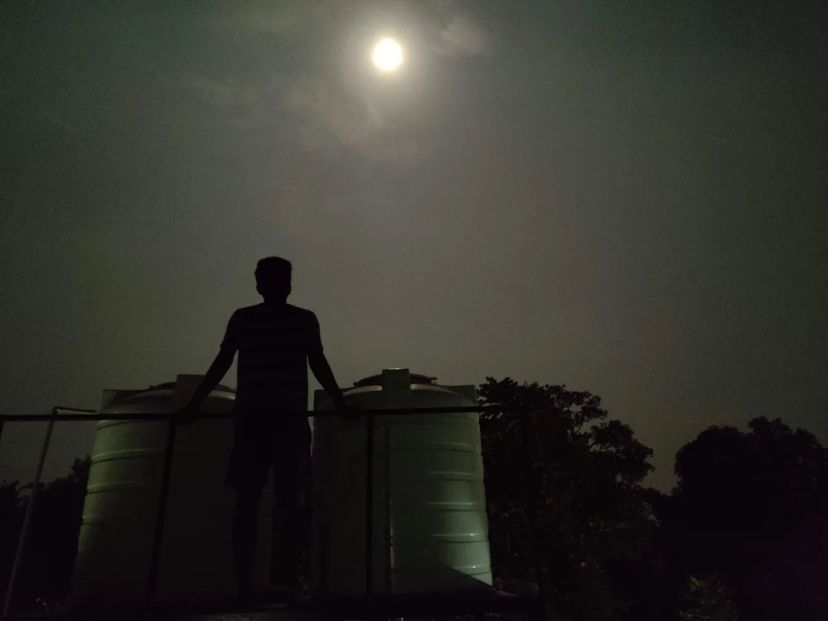 A silhouette of a person standing on a rooftop at night, leaning on a railing beside two large water tanks, gazing up at a…