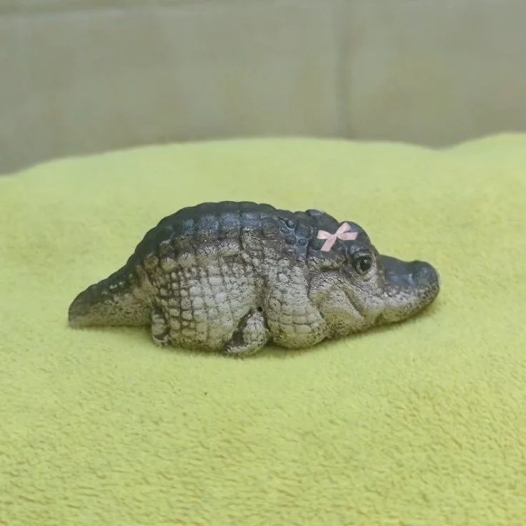 A small, baby alligator or crocodile rests on a bright yellow towel, with a tiny pink bow tied on its head. The image is…