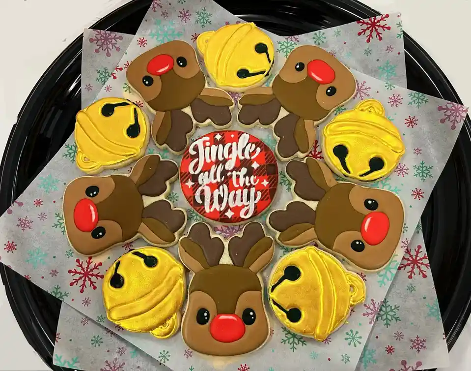 A tray of Christmas-themed sugar cookies shaped like reindeer with red noses and golden bells, arranged around a central…