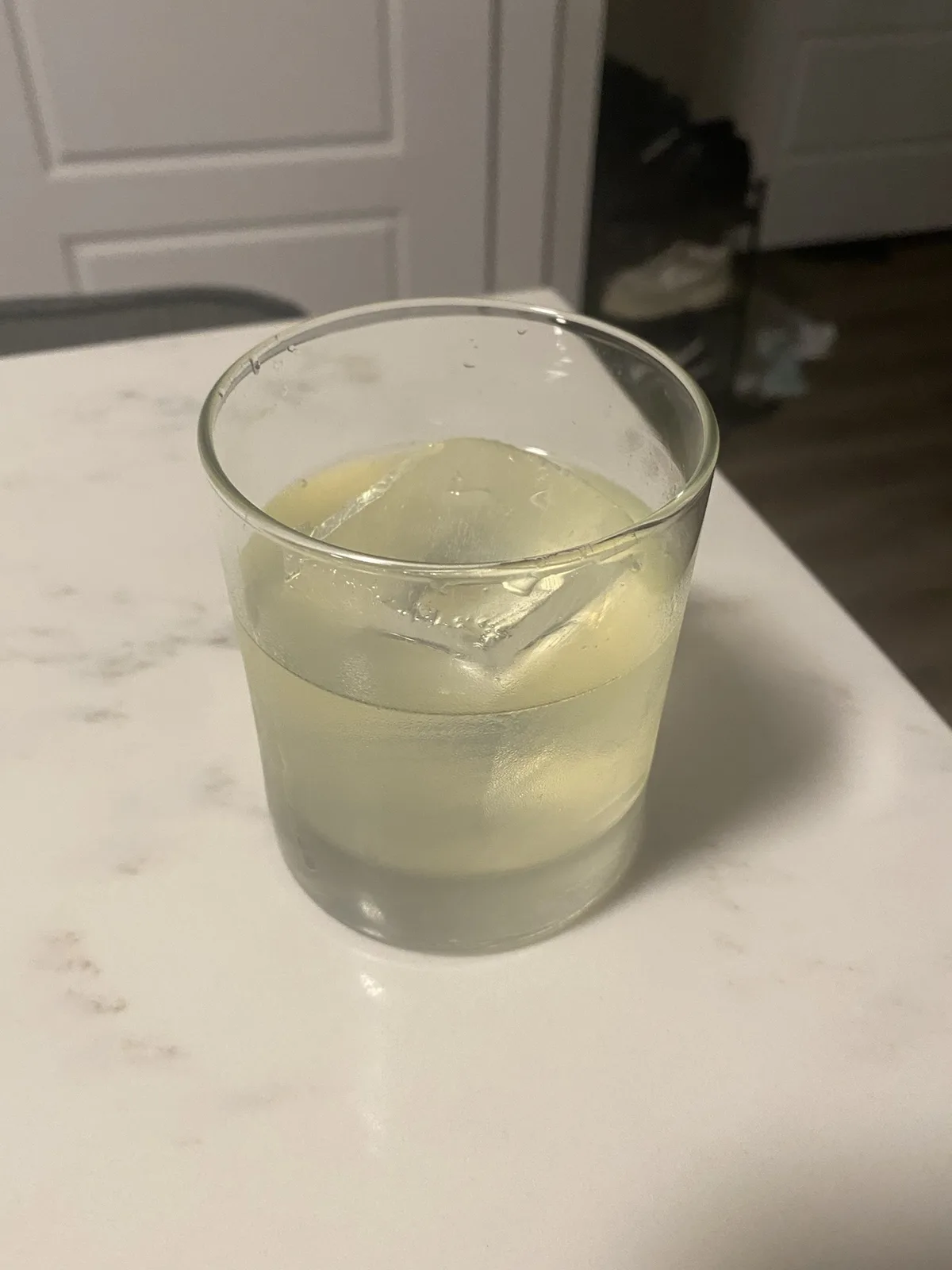 A clear glass containing a pale yellow liquid and a large ice cube sits on a white marble countertop, with a white door and…