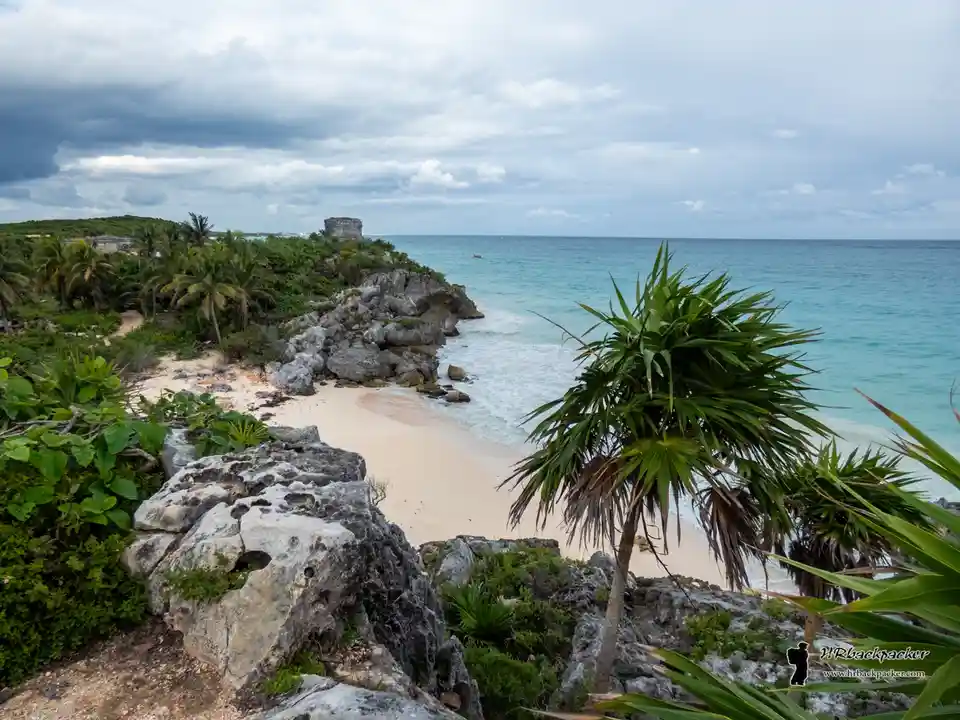 A scenic tropical beach photo with turquoise water, white sand, rocky cliffs, and palm trees under a cloudy sky. A stone…