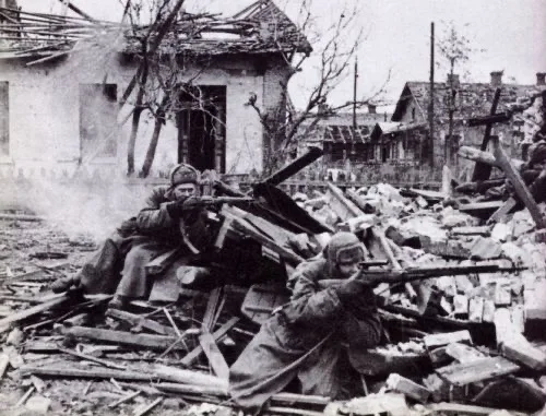 Two soldiers in winter gear are positioned behind rubble and debris in a destroyed urban area, aiming rifles. The background…
