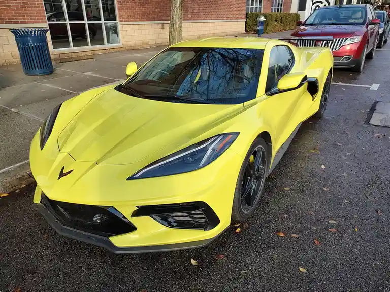A bright yellow Chevrolet Corvette sports car is parked in a public lot next to a red SUV, with a brick building and blue…
