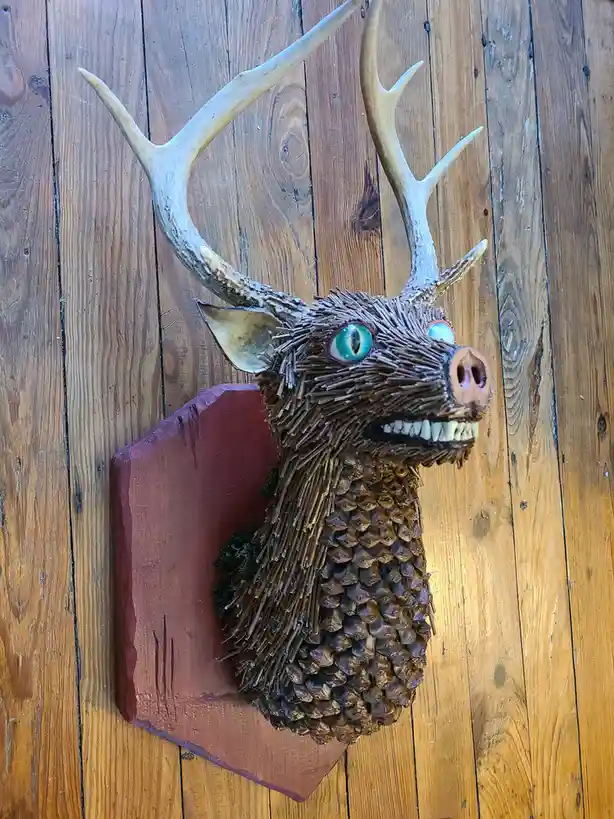 A taxidermy-style deer head mount is mounted on a wooden wall, but it has been humorously altered to have a pig snout, large…