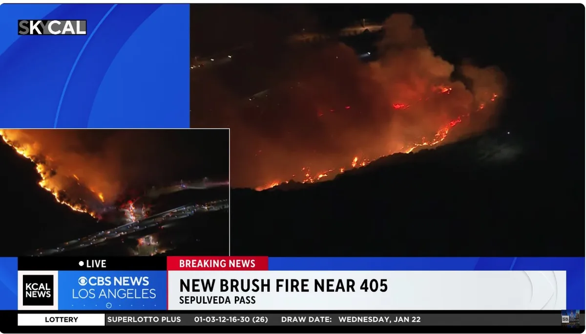 A news broadcast image shows a large brush fire at night, with flames and smoke visible on a hillside near a highway. The…