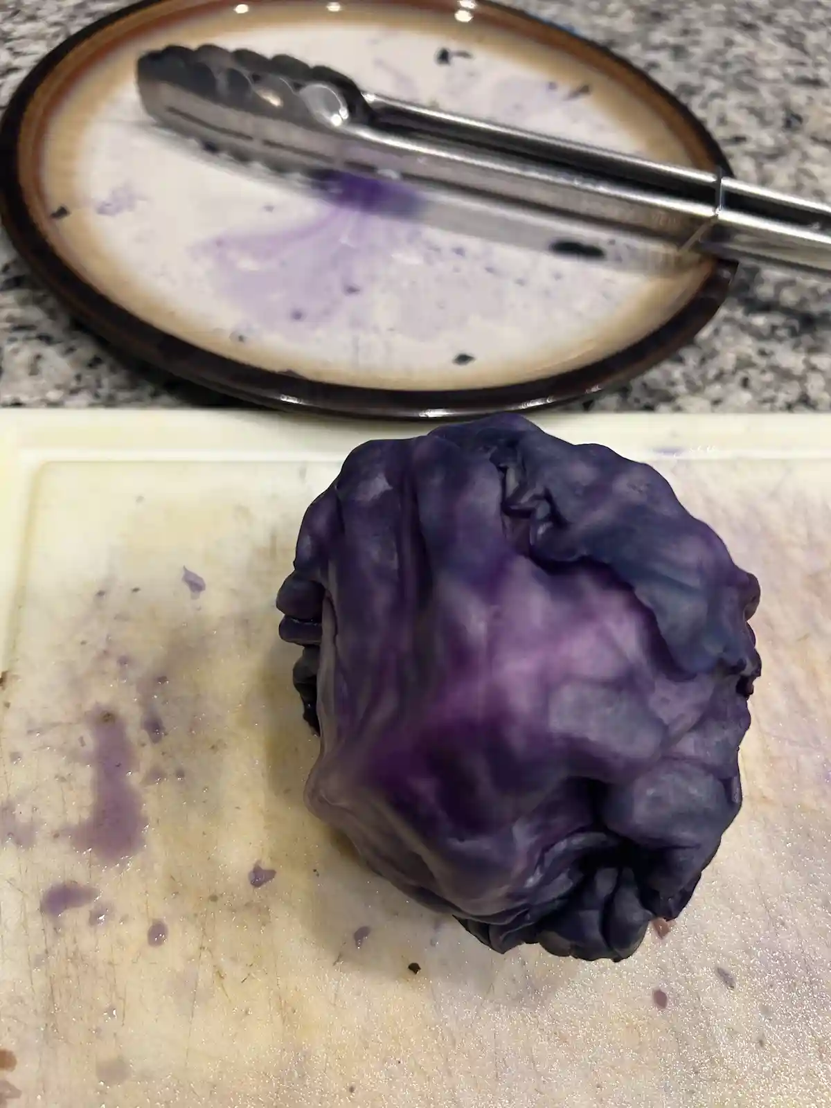 A whole purple cabbage sits on a white cutting board, with purple juice stains on the board. Behind it, a plate with metal…