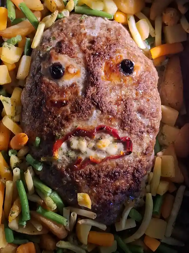 A cooked meatloaf shaped like a human face, with two black olives as eyes, a red pepper slice forming a mouth with melted…