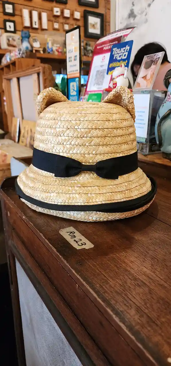A woven straw hat with two small ears on top and a black ribbon bow sits on a wooden counter in a shop. A small price tag…