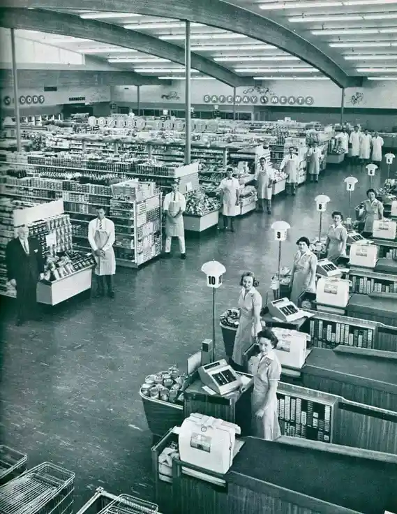 A black-and-white photograph of a spacious, mid-20th-century Safeway supermarket interior, showing aisles stocked with…