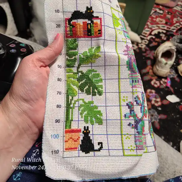 A hand holds up a cross-stitch project on white fabric with a grid, showing a partially completed design of two black cats…