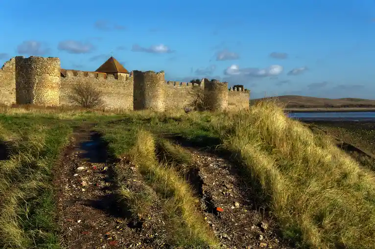 A scenic photo of a historic stone castle ruin with crenellated walls and a small tower under a blue sky with scattered…