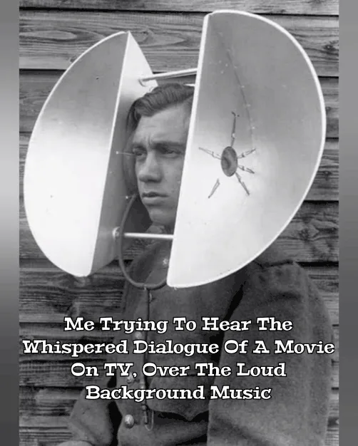 A black-and-white photo shows a man wearing a large, vintage, horn-style loudspeaker over his head, looking intently…