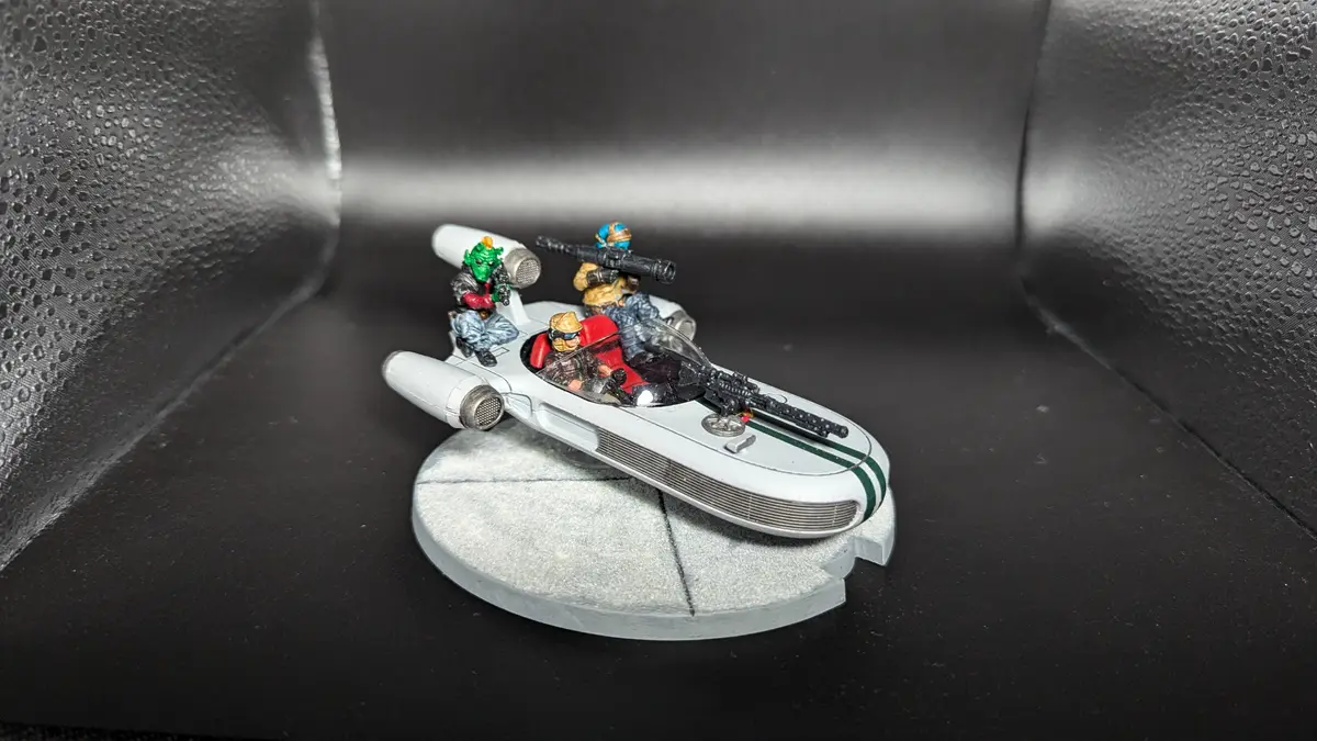 A painted miniature spaceship with four armed figures aboard sits on a grey base, placed on a black textured surface. The…