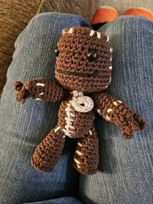 A handmade brown crocheted plush toy resembling a football, complete with white stitching and black button eyes, sits on a…