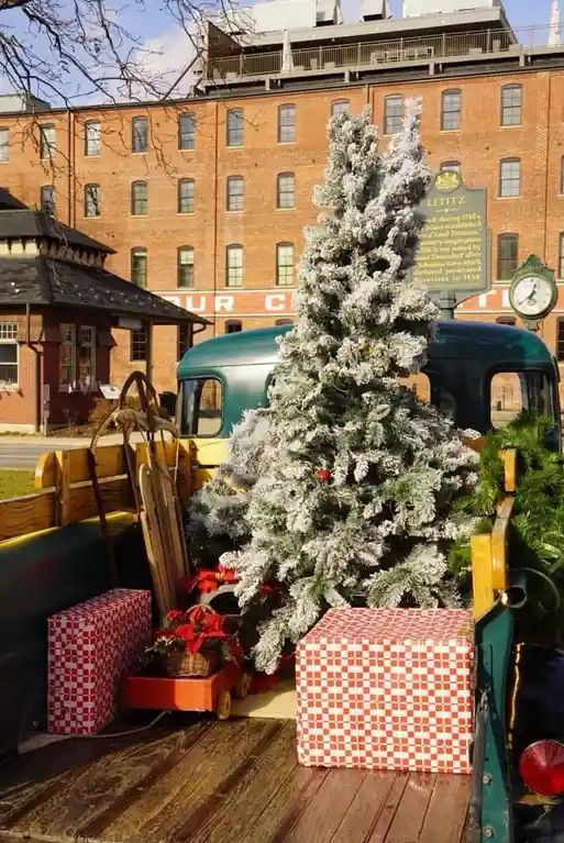 A flocked Christmas tree stands in the bed of a vintage green truck, surrounded by red-and-white checkered gift boxes and…