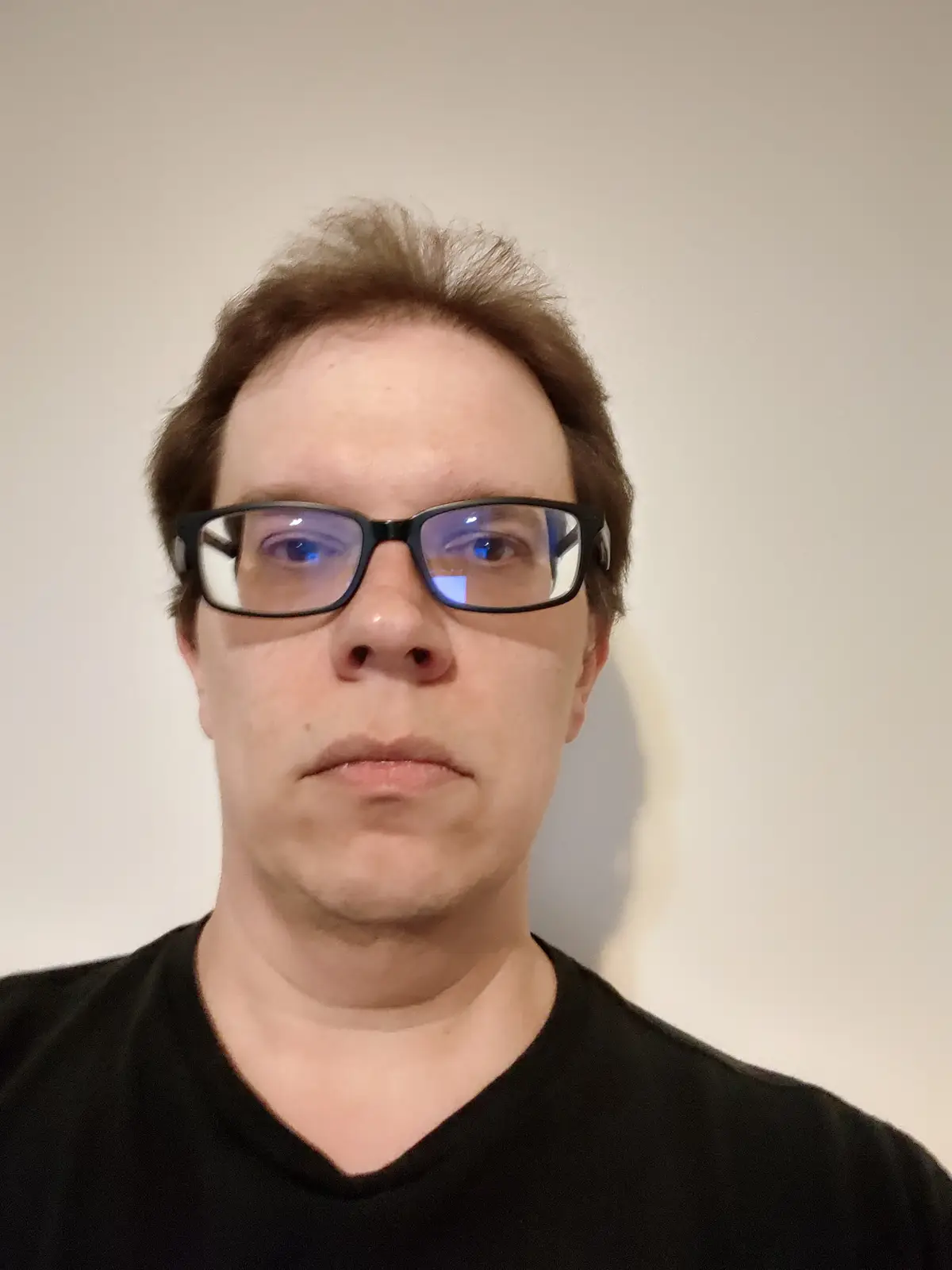 A close-up photo of a man with brown hair and black-rimmed glasses, wearing a black t-shirt, staring directly at the camera…