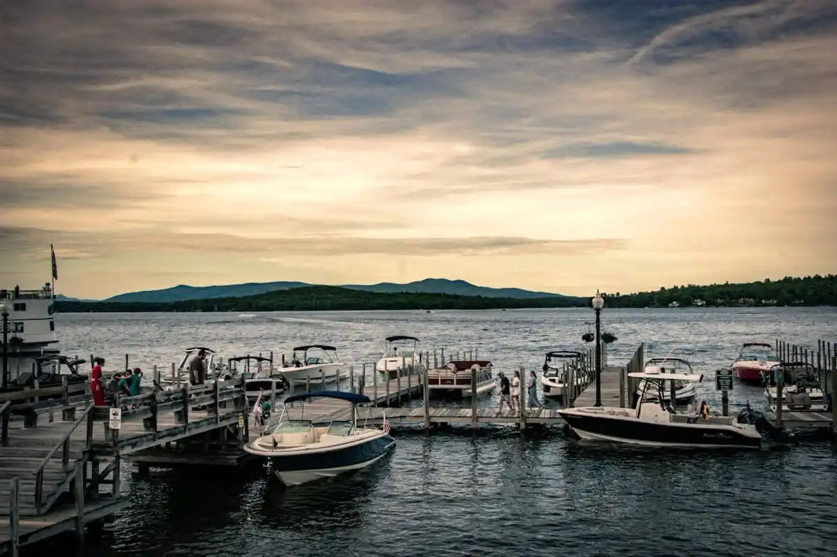 A scenic photo of a wooden dock with several boats moored at a lake, people walking along the pier, and mountains visible in…