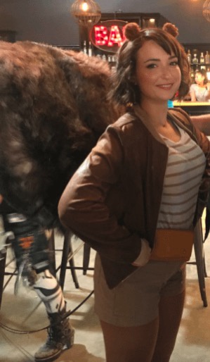 A woman with small brown bear ears on her head smiles at the camera while standing next to a large, furry bear costume in…