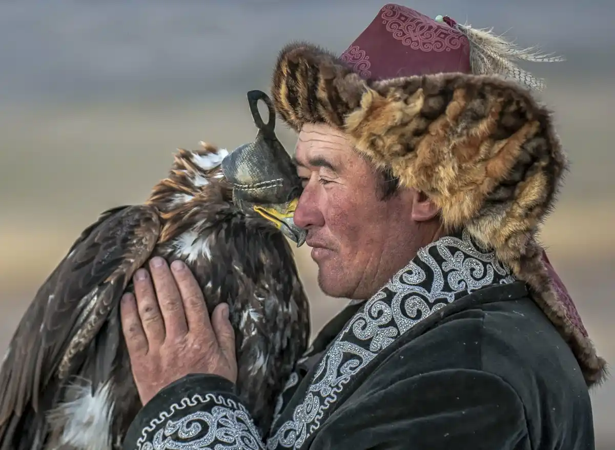 A man in traditional Central Asian attire, wearing a fur-lined hat and embroidered robe, gently holds a large golden eagle…