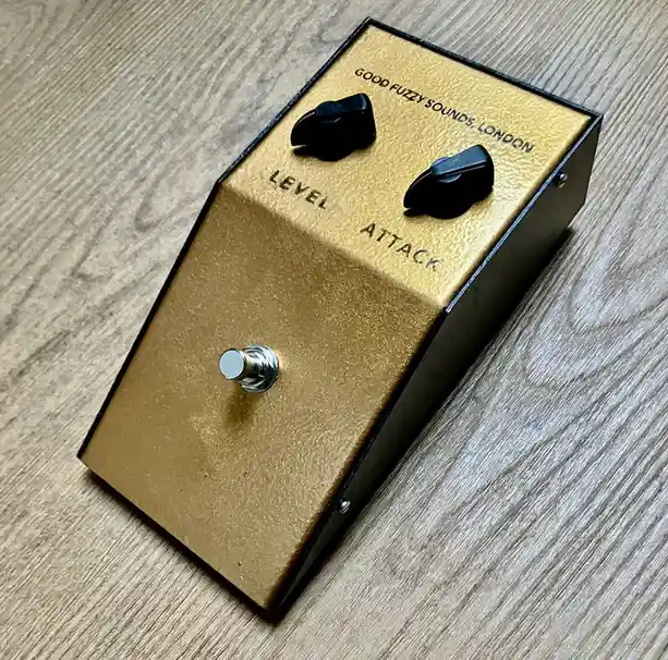 A gold-colored guitar effects pedal with black knobs labeled 'LEVEL' and 'ATTACK', and a silver footswitch, resting on a…