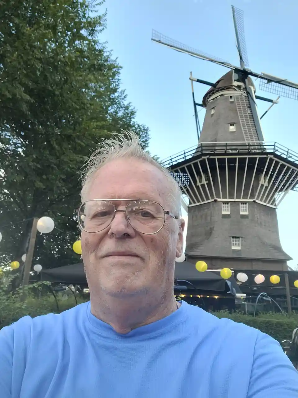 A selfie of a middle-aged man with glasses and a blue shirt, standing in front of a large traditional Dutch windmill under a…
