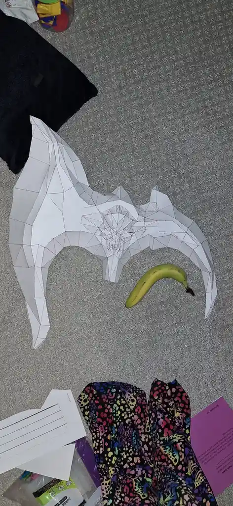 A large, white, geometrically patterned paper cutout of a bat lies on a gray carpet, with a single yellow banana placed…
