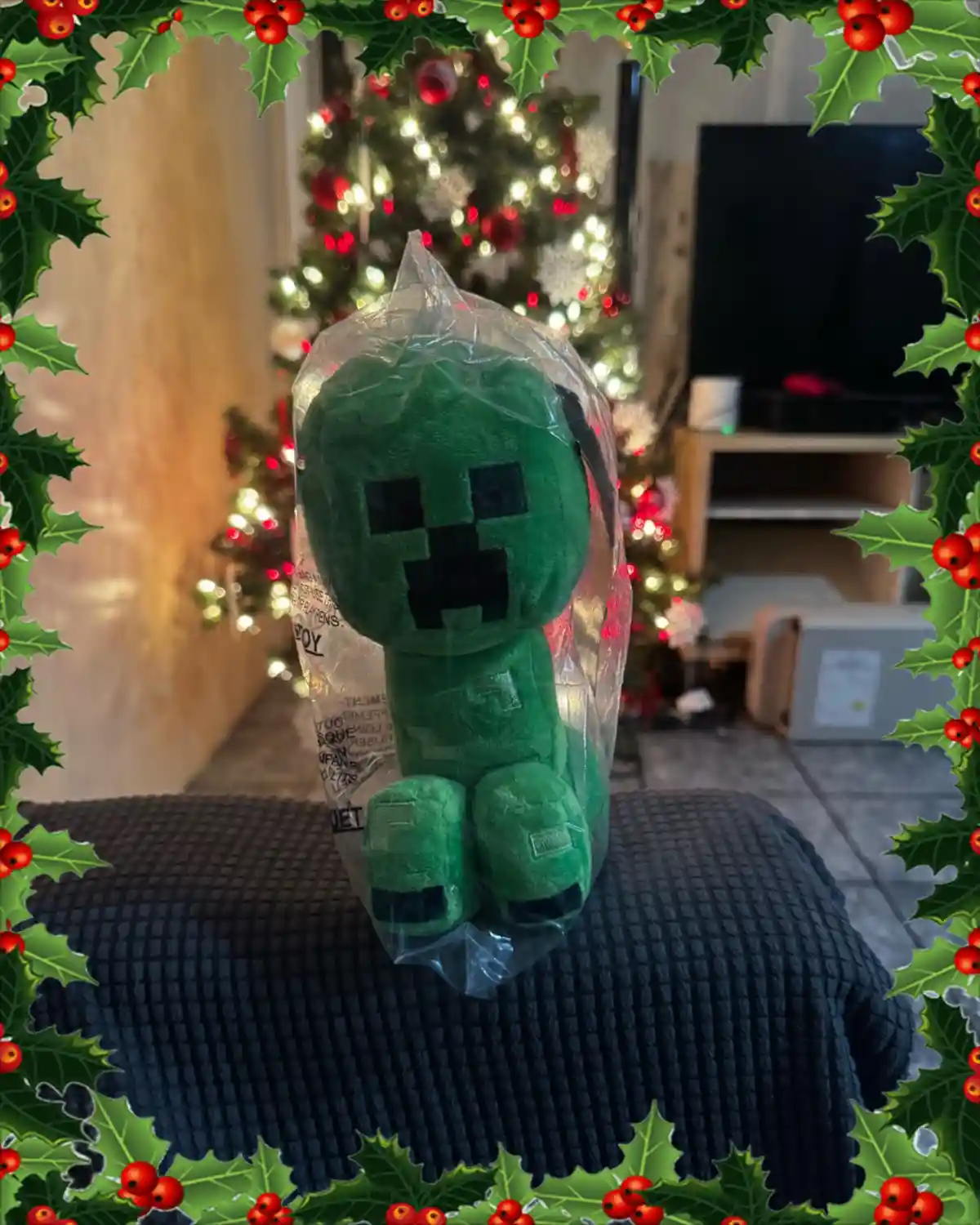 A green Minecraft creeper plush toy, still in its clear plastic packaging, sits on a dark textured blanket. The background…